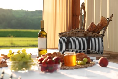 Photo of Romantic date. Wicker basket, glass of wine and snacks for picnic on white blanket outdoors