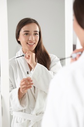 Photo of Attractive young woman applying oil onto her eyelashes near mirror indoors