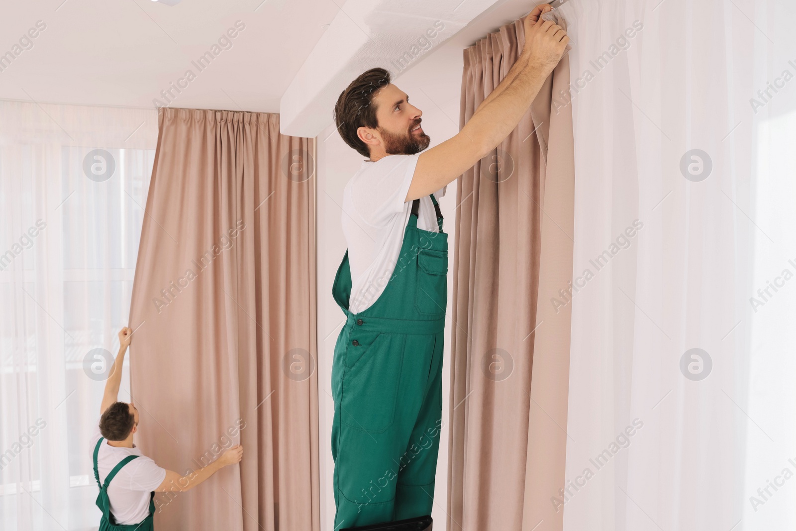 Photo of Workers in uniform hanging window curtain indoors