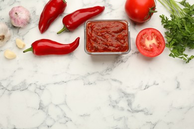 Photo of Delicious adjika sauce in bowl and ingredients on white marble table, flat lay. Space for text