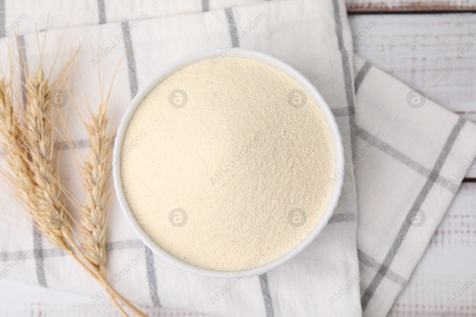 Photo of Uncooked organic semolina in bowl and spikelets on white wooden table, top view