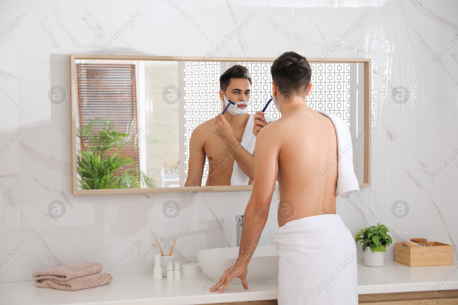 Photo of Handsome young man shaving with razor near mirror in bathroom