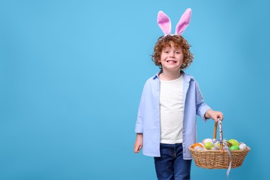 Photo of Happy boy in cute bunny ears headband holding wicker basket with Easter eggs on light blue background. Space for text