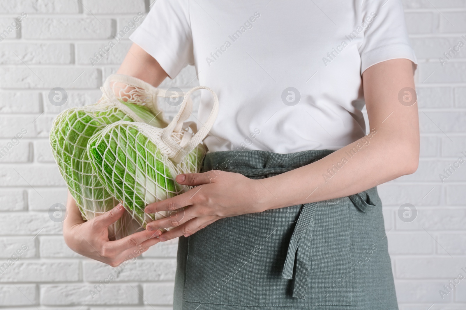 Photo of Woman holding string bag with fresh Chinese cabbages near white brick wall, closeup