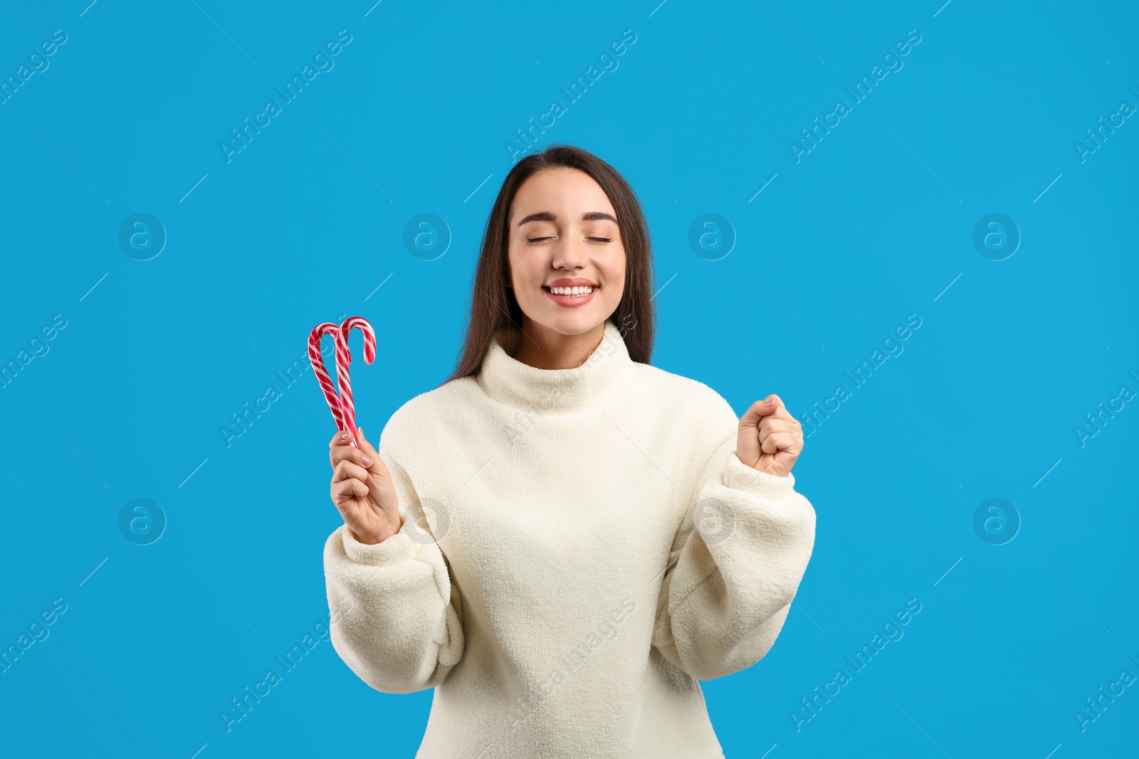 Photo of Young woman in beige sweater holding candy canes on blue background. Celebrating Christmas