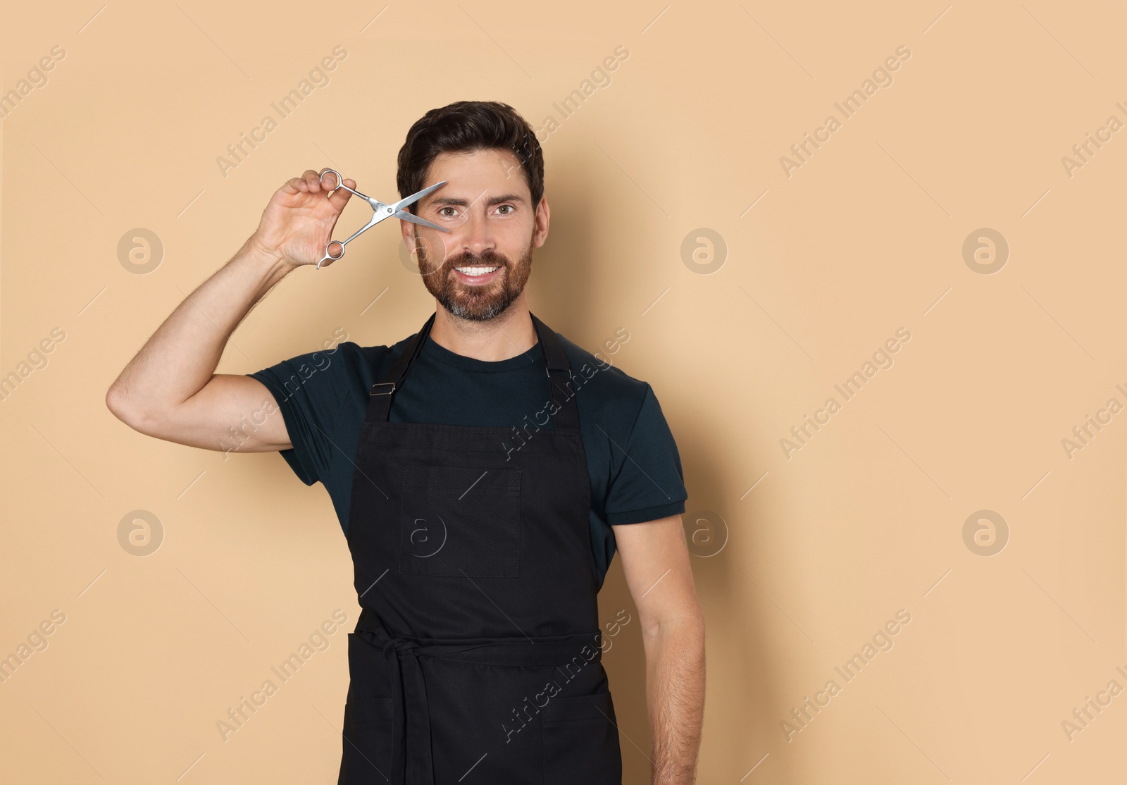 Photo of Smiling hairdresser in apron holding scissors on light brown background, space for text