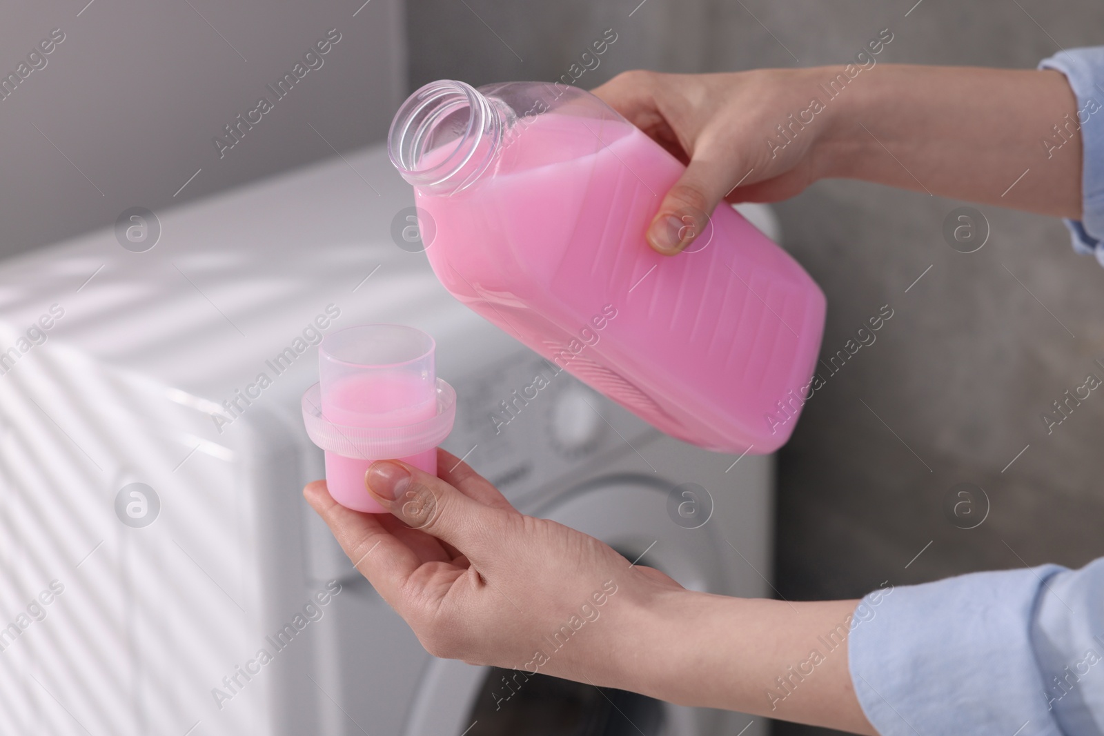 Photo of Woman pouring fabric softener from bottle into cap for washing clothes indoors, closeup