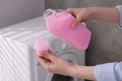 Photo of Woman pouring fabric softener from bottle into cap for washing clothes indoors, closeup