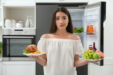Photo of Woman choosing between fruits and burger with French fries near refrigerator in kitchen
