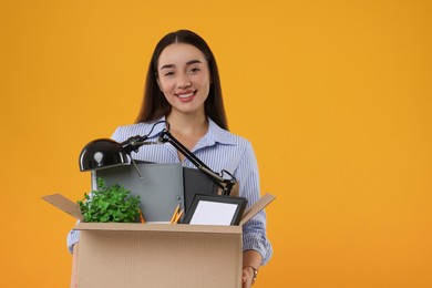 Happy unemployed woman holding box with personal office belongings on orange background, space for text