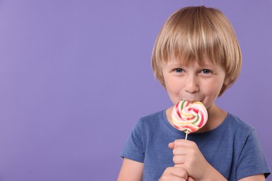 Photo of Happy little boy licking colorful lollipop swirl on violet background, space for text