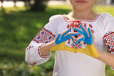 Photo of Little girl making heart with her hands painted in Ukrainian flag colors outdoors, closeup and space for text. Love Ukraine concept