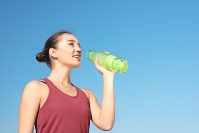 Photo of Young sporty woman drinking water from bottle against blue sky on sunny day. Space for text