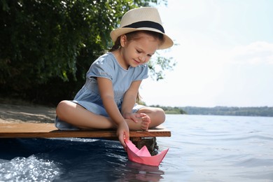 Photo of Cute little girl playing with paper boat on wooden pier near river