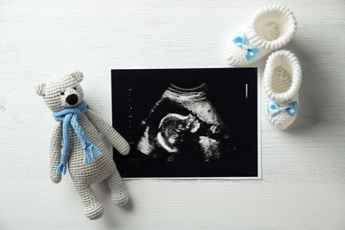 Photo of Ultrasound photo of baby, toy and knitted boots on wooden background, flat lay