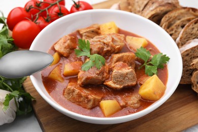 Photo of Delicious goulash in bowl, bread and ingredients on white table, closeup