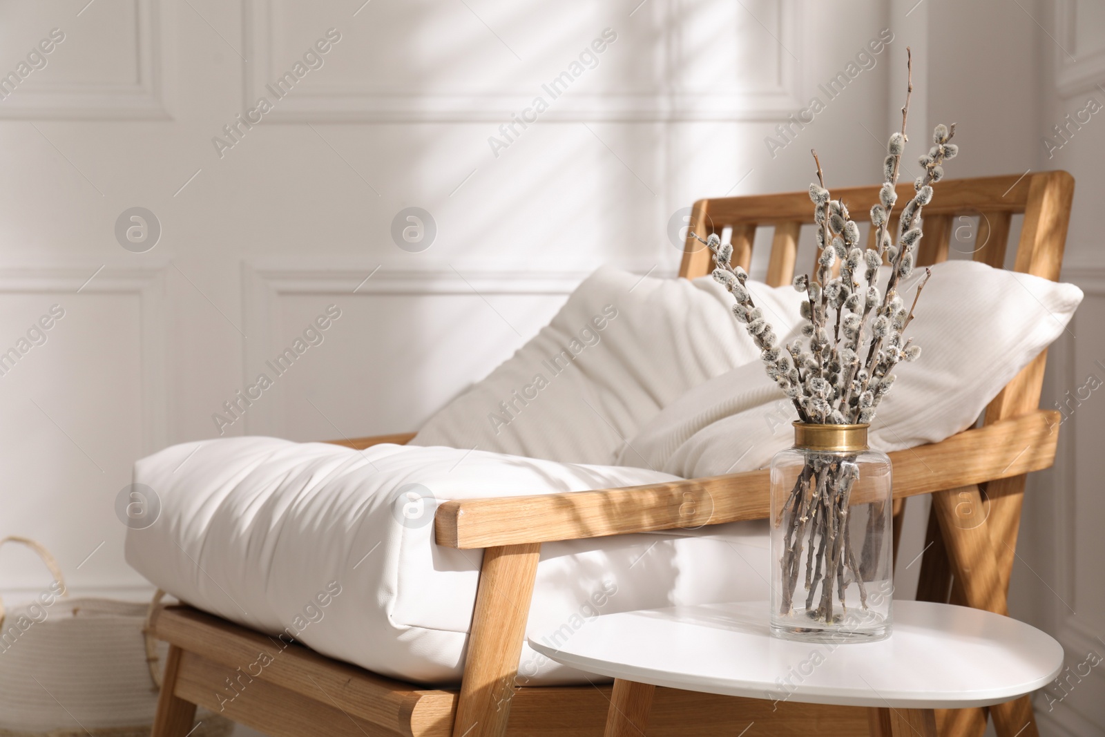 Photo of Glass vase with pussy willow tree branches on table near armchair indoors, space for text