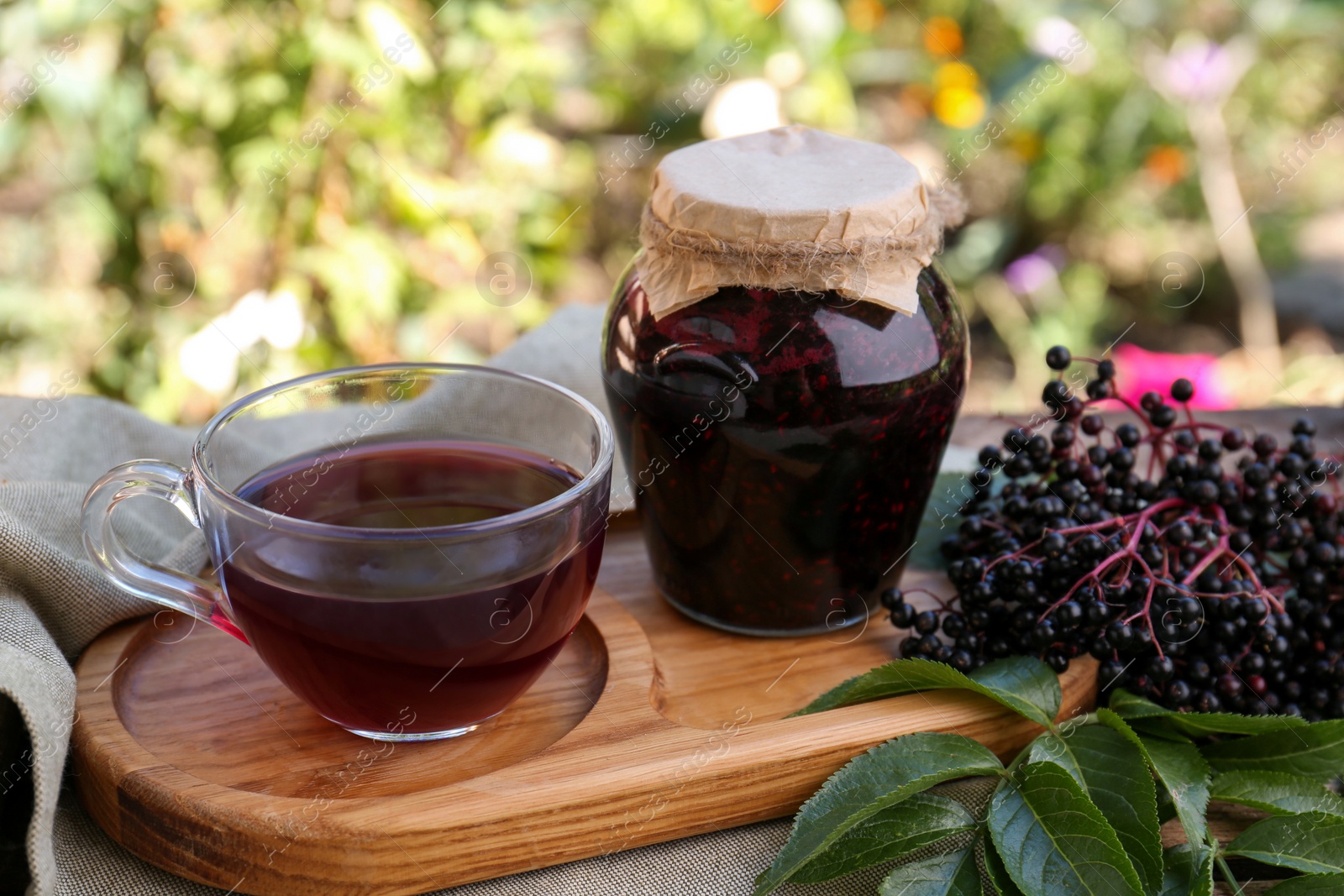 Photo of Elderberry jam, glass cup of tea and Sambucus berries on table outdoors