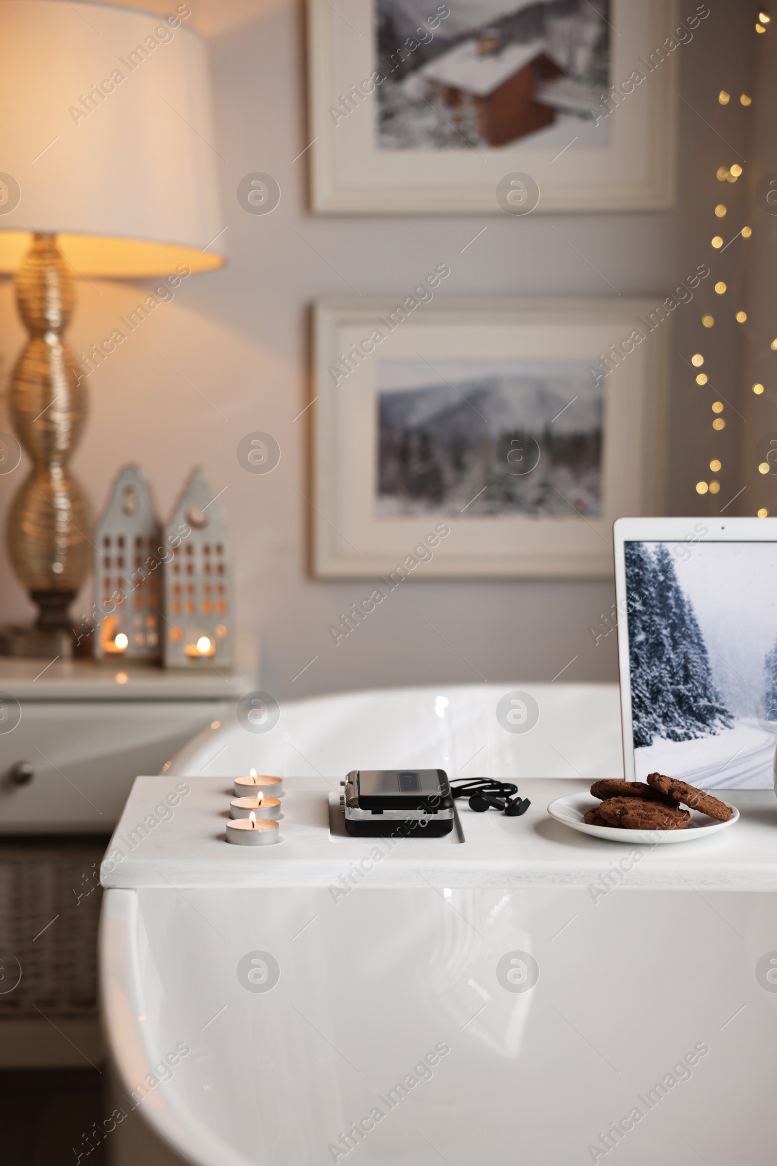 Photo of White wooden tray with tablet, cassette player, cookies and burning candles on bathtub in bathroom