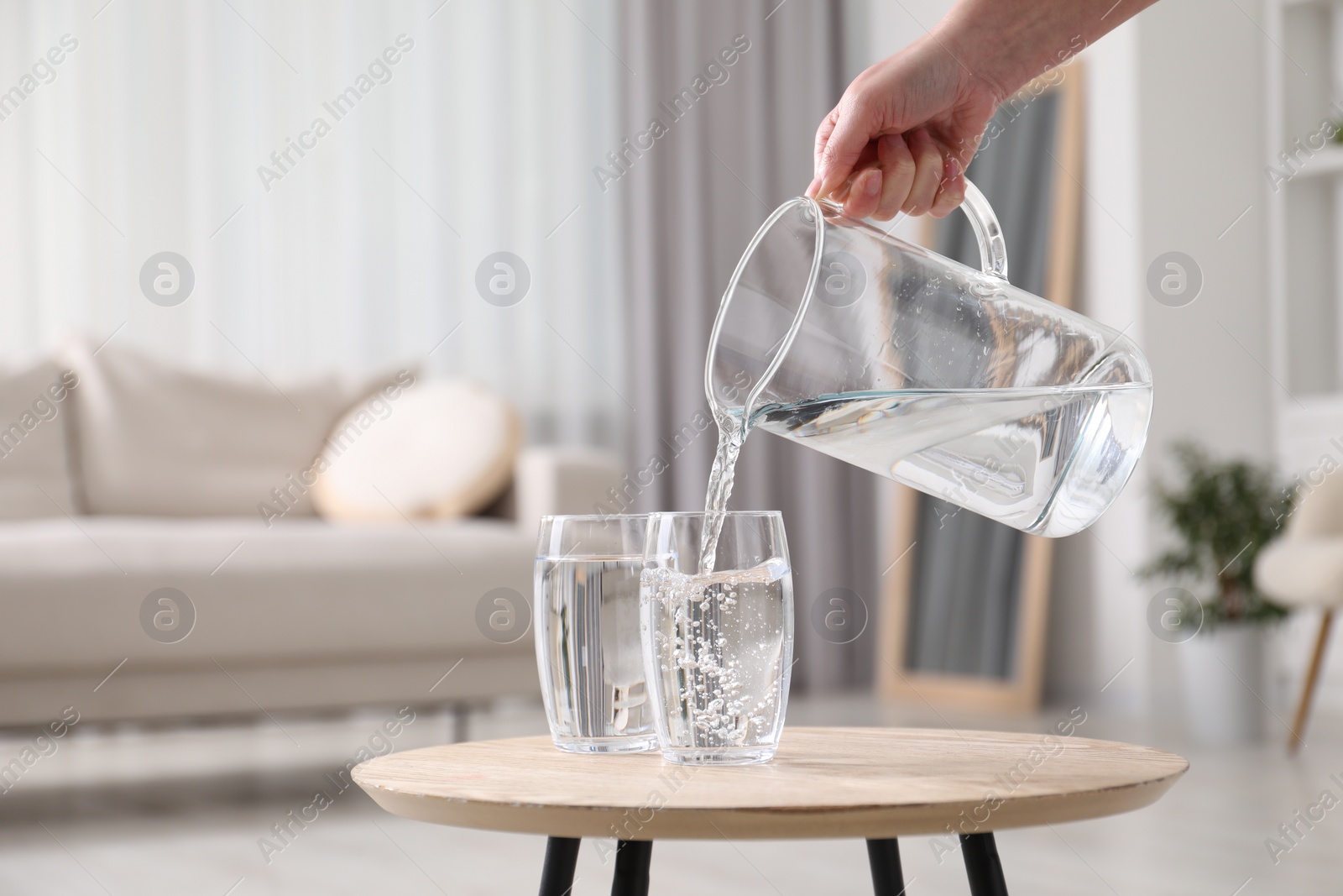 Photo of Woman pouring fresh water from jug into glass at wooden table indoors, closeup. Space for text