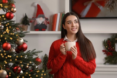 Photo of Smiling woman holding cup of hot drink near Christmas tree at home