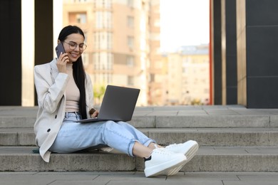 Happy young woman using modern laptop and talking on smartphone outdoors. Space for text