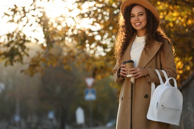 Photo of Portrait of beautiful African-American woman with stylish white backpack and hot drink on city street. Space for text