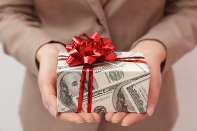 Photo of Woman holding gift box wrapped in dollars on light background, closeup