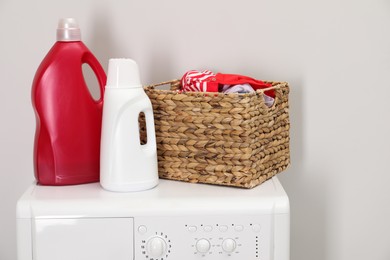 Photo of Baby clothes in wicker basket and laundry detergents on washing machine near light wall