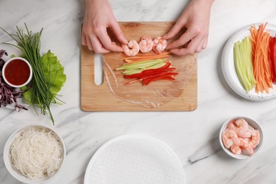 Photo of Making delicious spring rolls. Woman wrapping fresh vegetables and shrimps into rice paper at white marble table, top view