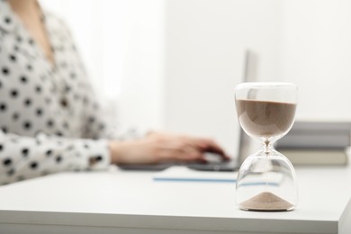 Photo of Hourglass with flowing sand on desk. Woman using laptop indoors, selective focus