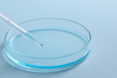 Photo of Dripping liquid from pipette into petri dish at light blue background, closeup. Laboratory analysis