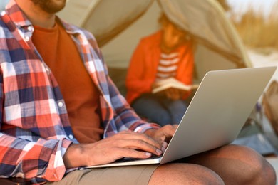 Photo of Man using laptop outdoors while his girlfriend reading book in camping tent, closeup