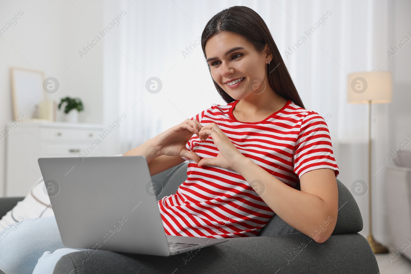 Photo of Happy young woman having video chat via laptop and making heart with hands on sofa in living room