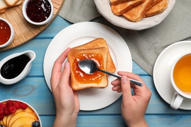 Photo of Woman spreading apricot jam onto tasty toast at light blue wooden table, top view