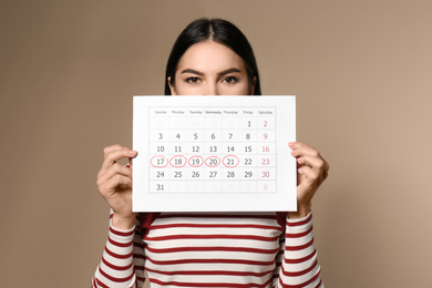 Photo of Young woman holding calendar with marked menstrual cycle days on beige background