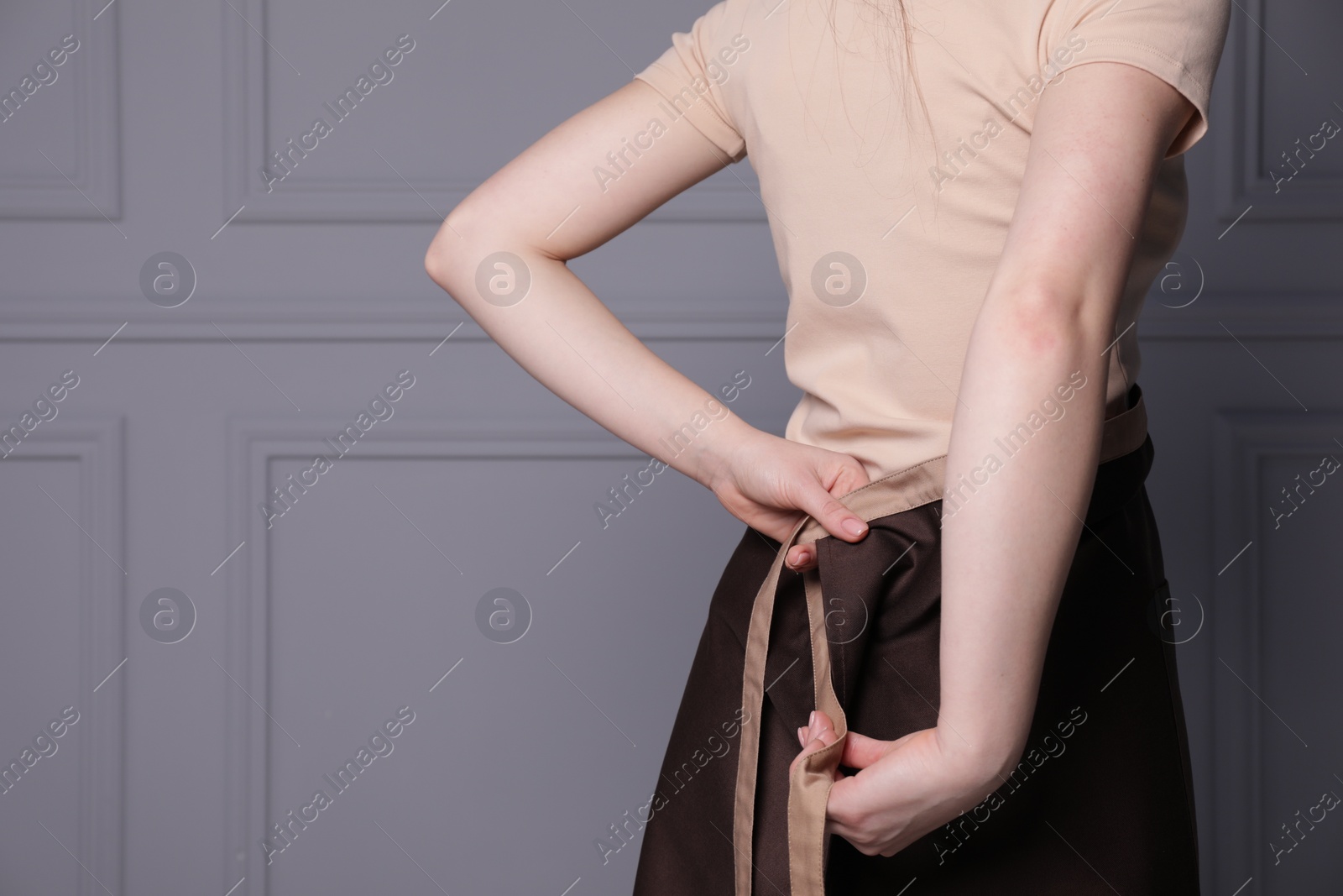 Photo of Woman putting on brown apron against grey wall, closeup. Space for text