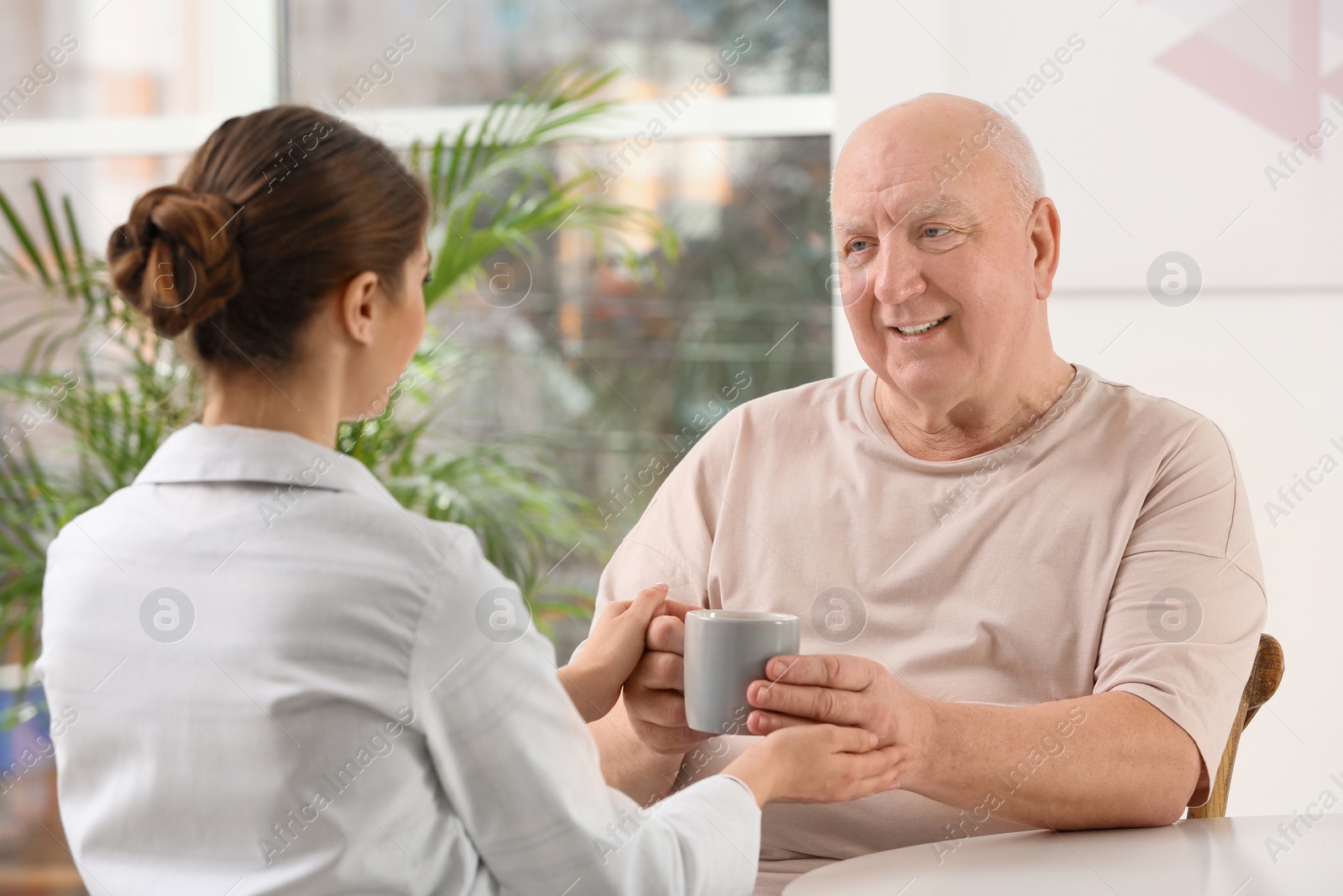 Photo of Nurse giving cup of tea to elderly man indoors. Assisting senior generation