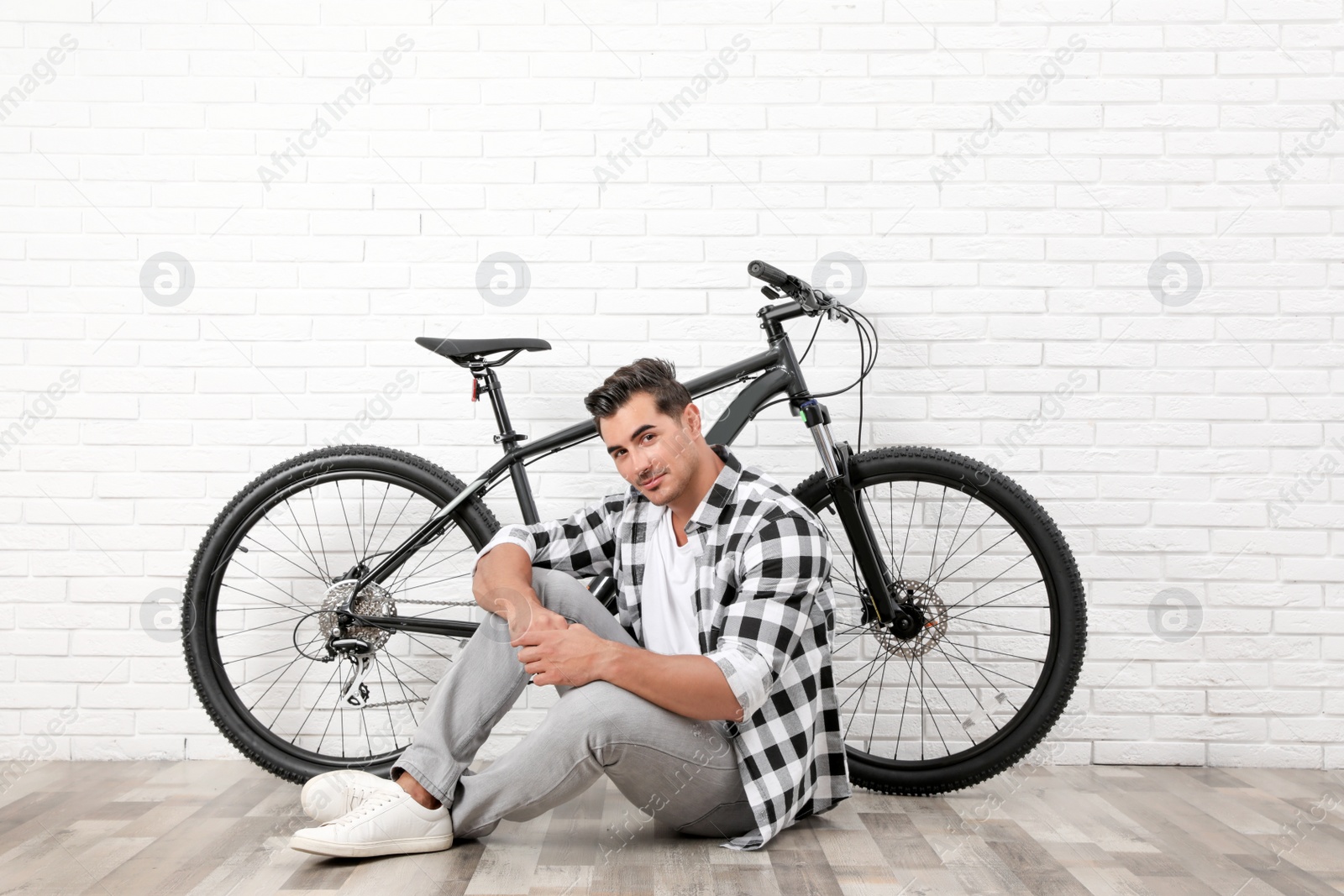 Photo of Handsome young man with modern bicycle near white brick wall indoors