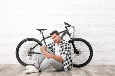 Photo of Handsome young man with modern bicycle near white brick wall indoors