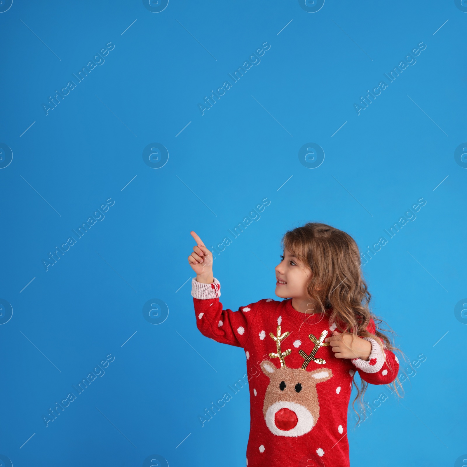 Photo of Cute little girl in red Christmas sweater pointing against blue background. Space for text
