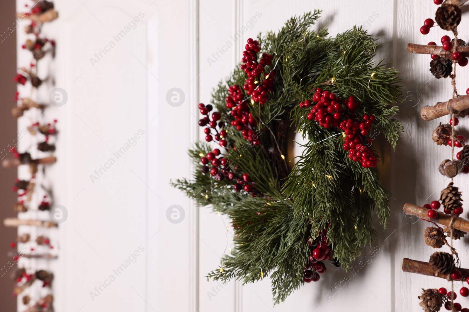 Photo of Beautiful Christmas wreath with red berries and fairy lights hanging on white door, space for text