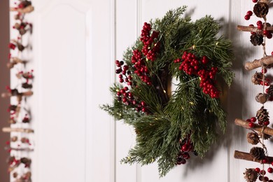 Photo of Beautiful Christmas wreath with red berries and fairy lights hanging on white door, space for text