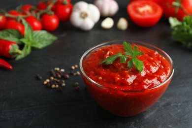 Photo of Delicious adjika sauce with parsley in glass bowl and ingredients on black table, closeup