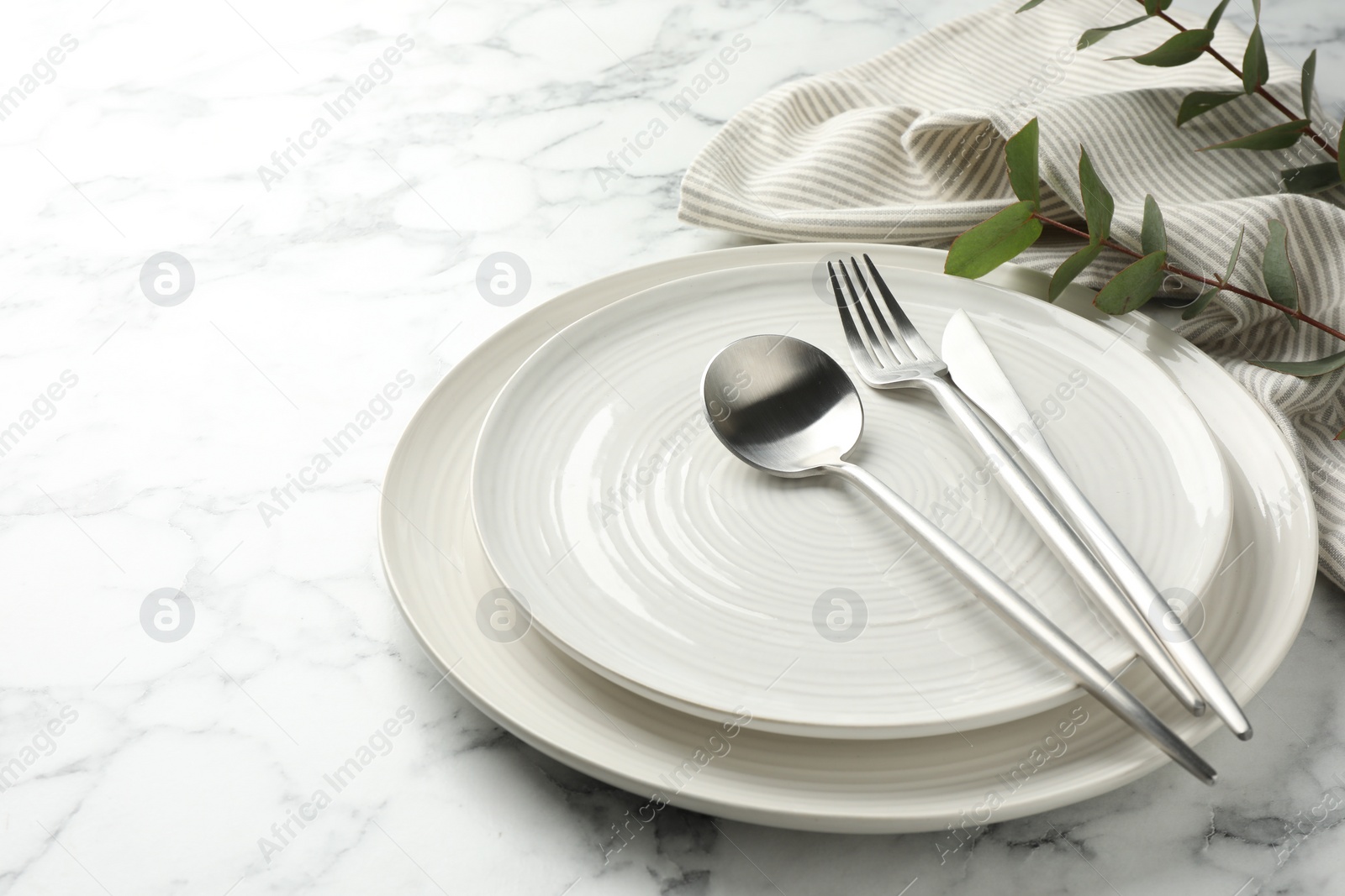 Photo of Stylish setting with cutlery, napkin, eucalyptus branches and plates on white marble table, closeup. Space for text