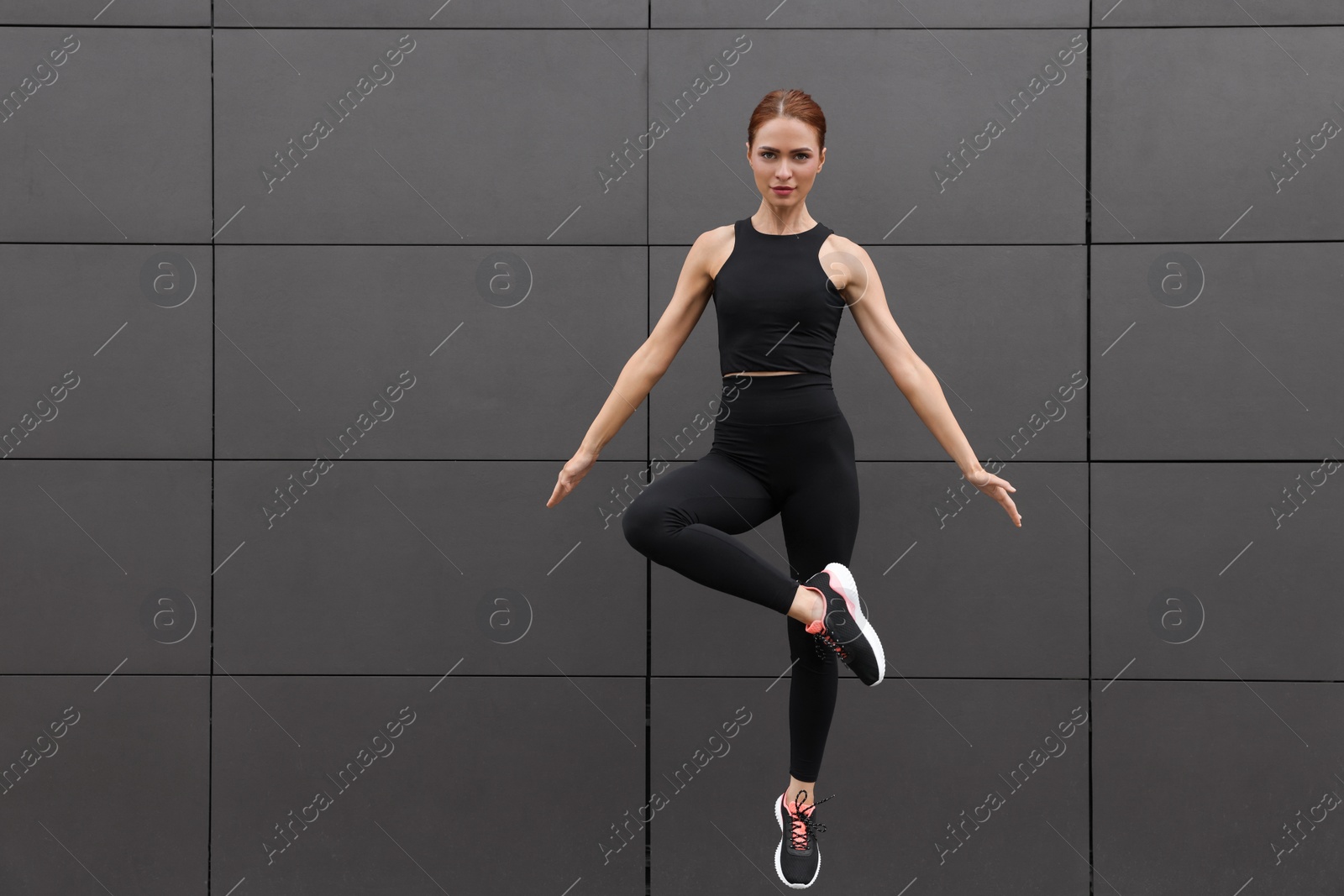 Photo of Beautiful woman in gym clothes jumping near dark grey wall on street, space for text