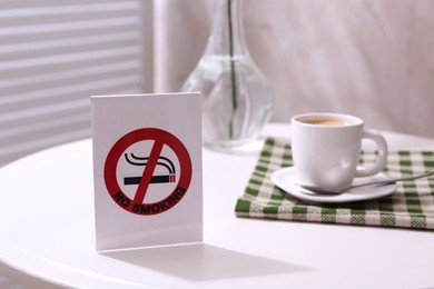 Photo of No Smoking sign and cup of coffee on white table indoors