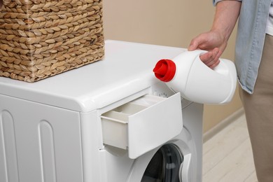 Photo of Man pouring fabric softener from bottle into washing machine indoors, closeup