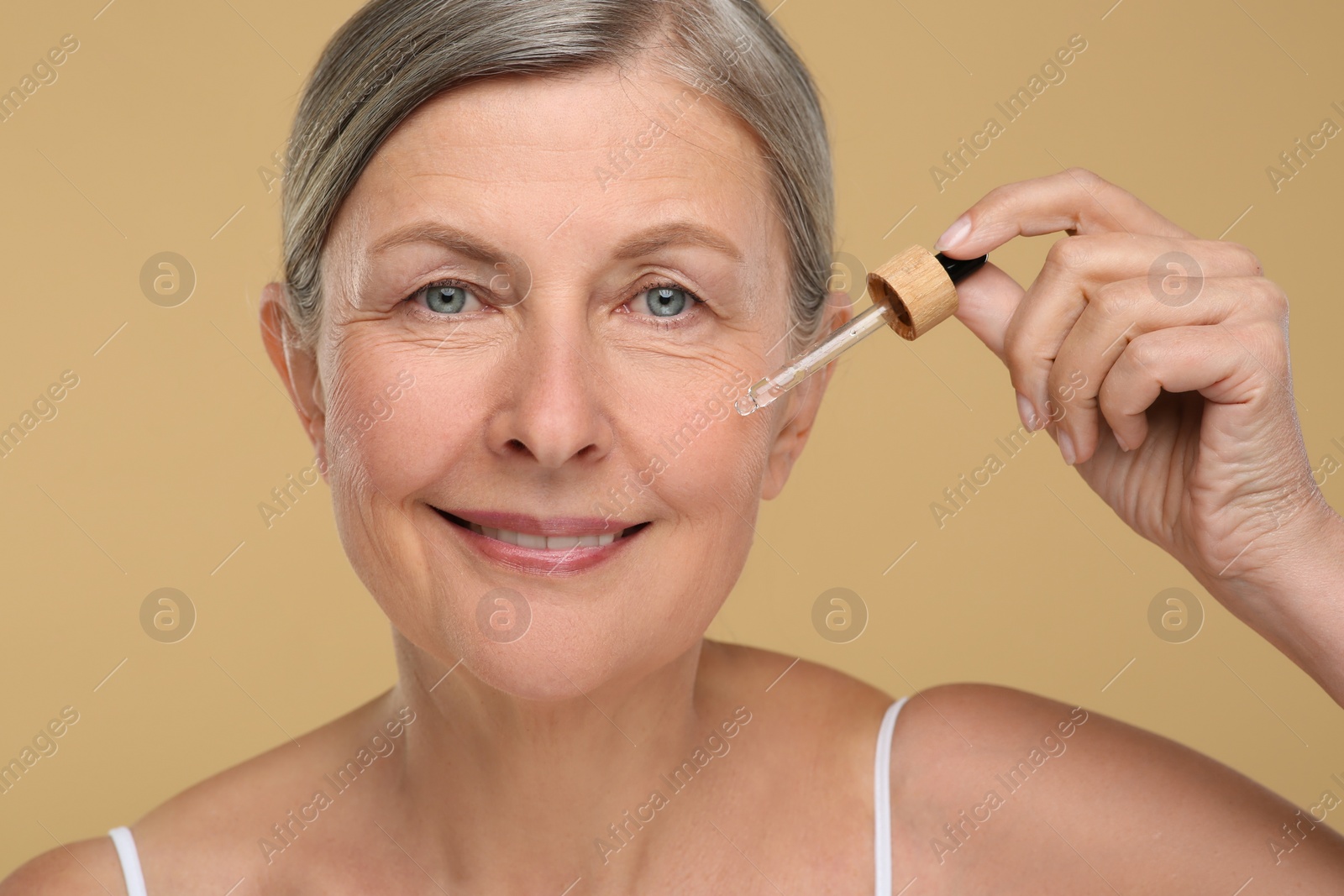 Photo of Senior woman applying cosmetic product on her aging skin against beige background. Rejuvenation treatment