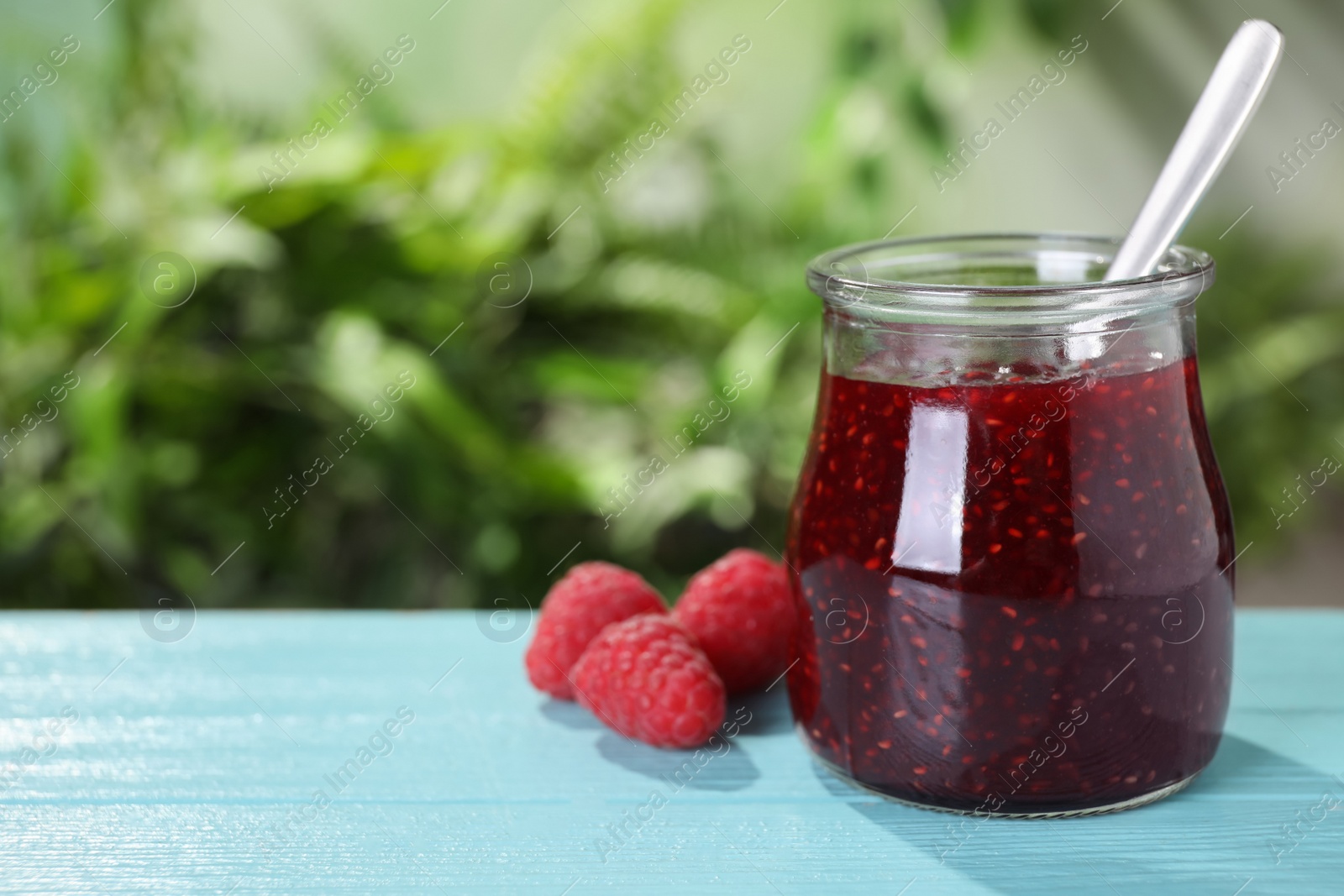 Photo of Delicious jam in glass jar and fresh raspberries on blue wooden table. Space for text